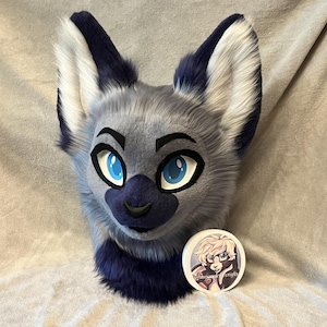 May include: A grey and dark blue faux fur animal head mask featuring large pointed ears with white inner fur, bright blue eyes with black outlines, and a dark blue nose. A circular white badge with an illustrated character and the text @moondelcrafts is placed in front.