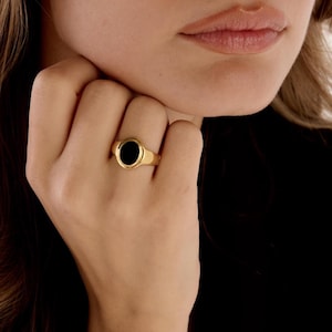 May include: A gold ring with a black oval stone. The ring is on a hand with long blonde hair and a black top in the background. The ring is a classic signet style.