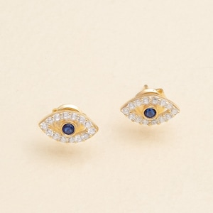 May include: A pair of gold earrings with a blue and white evil eye design. The earrings are small and delicate.