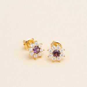 May include: A pair of gold stud earrings with a purple gemstone surrounded by smaller white gemstones in a flower shape.