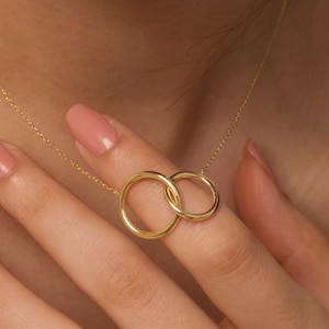 May include: A gold chain necklace with a pendant featuring two interlocking circles. The necklace is worn by a person with light skin and pink nail polish.