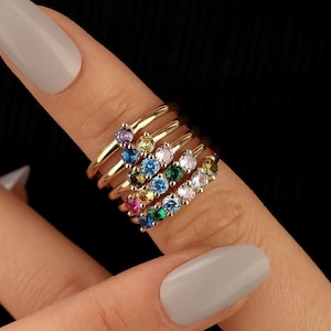 May include: A stack of gold-colored rings, each adorned with a row of colorful gemstones. The rings are displayed on a finger against a dark background. The gemstones include purple, yellow, blue, green, and white.