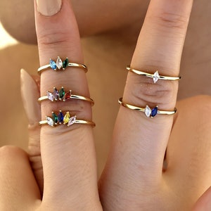 May include: A set of five gold rings with colorful marquise-shaped gemstones. Each ring features a single gemstone in a different color, including green, red, blue, yellow, and clear.