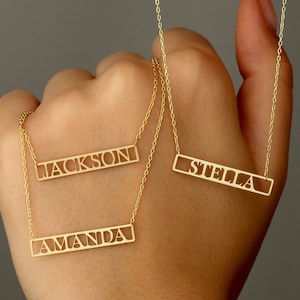 May include: Three gold necklaces with personalized nameplates. The nameplates are rectangular and have the names "Amanda", "Jackson", and "Stella" cut out in the metal.