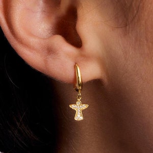 May include: Gold hoop earrings with a dangling angel charm. The angel is adorned with small, clear cubic zirconia stones.