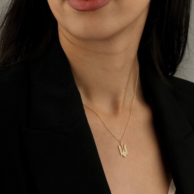 May include: A gold necklace with a pendant featuring the Ukrainian trident symbol. The necklace is worn by a person with long dark hair and a black blazer.