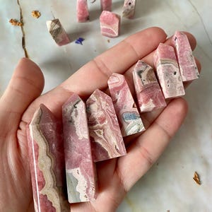 May include: A selection of pink and white gemstone towers with diverse patterns and shapes. The crystals are held in a hand, with others scattered on a light surface. The stones have a polished finish and display natural veining. The crystals are measured in cm.