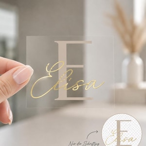 Könnte beinhalten: Ein quadratischer, transparenter Aufkleber mit einem großen, hellbeigen Großbuchstaben E, überlagert vom Namen Elisa in goldener Schreibschrift. Ein kreisförmiger Einschub zeigt das Design auf transparentem Hintergrund mit deutschem Text.