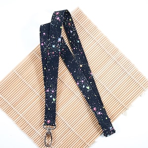 May include: A black lanyard with a colourful splatter pattern and a silver swivel clip. The lanyard is lying on a bamboo mat.