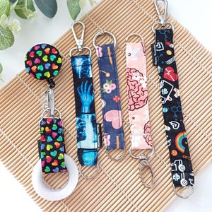 May include: A collection of five lanyards and badge reels with various patterns. Designs include colorful hearts, an X-ray of a hand, floral patterns, a brain illustration, and science-themed graphics. Each lanyard has a metal clasp.