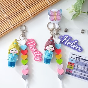 Könnte beinhalten: Zwei ausziehbare Ausweishalter mit Cartoon-Figuren in blauer Arbeitskleidung, bunten herzförmigen Perlen und Namensschildern mit der Aufschrift Verity und Helen. Ein Halter hat einen Schmetterling, dazu eine Referenzkarte für Blutabnahmen.
