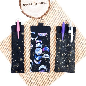 Könnte beinhalten: Drei schwarze Stifthalter mit verschiedenen Himmelsmustern. Einer hat goldene Sterne, ein anderer Mondphasen in Blau und Lila, und der dritte hat goldene Sterne. Jeder hat einen Stift und eine schwarze Schlaufe zur Befestigung.