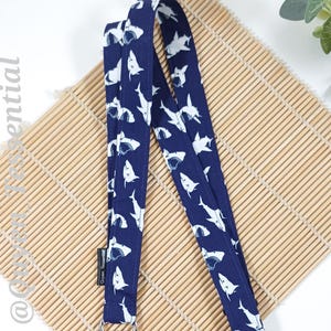 May include: A navy blue lanyard featuring a white shark design. The fabric lanyard has a silver-coloured metal clasp. It rests on a bamboo mat.