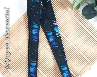 May include: Black lanyard featuring a celestial moon phase pattern in shades of blue and purple. The lanyard has a silver-colored metal clasp and is displayed on a bamboo mat. The text "@Quyen_Tessential" is visible on the left side.