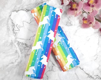 May include: Two rainbow-striped fabric car seat belt covers with white unicorn silhouettes and white stars.