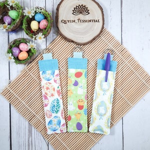 May include: Three Easter-themed pen holders with key rings. The holders feature colourful patterns of Easter eggs, bunnies, and chicks. Each holder has a light blue top and a silver key ring. The background includes decorative nests with Easter eggs.