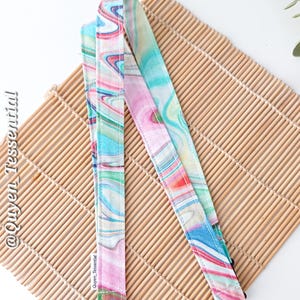 May include: A colorful lanyard with a marbled pattern in shades of pink, blue, and green. The lanyard has a silver-colored swivel clasp and is lying on a bamboo mat. The text "@Quyen Tessential" is printed on the lanyard.