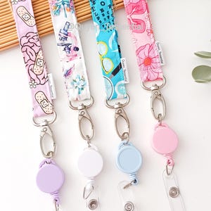 May include: Four patterned lanyards with silver clasps and retractable badge reels. Designs include bandages, flowers, eyeglasses, and a floral pattern. The reels are in purple, white, light blue, and pink. The lanyards are displayed on a light brown surface.