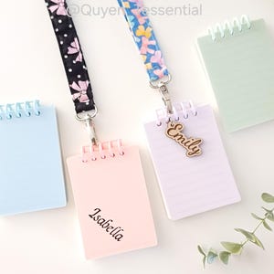 May include: Three small spiral notebooks with personalised name tags attached to lanyards. The notebooks are light blue, pink, and green. The name tags are made of wood and say "Emily", "Isabella", and "Emily".
