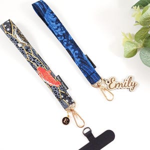May include: Two fabric phone straps with gold hardware and a wooden name tag that says "Emily". One strap has a blue and white pattern and the other has a grey and gold pattern with red fish.