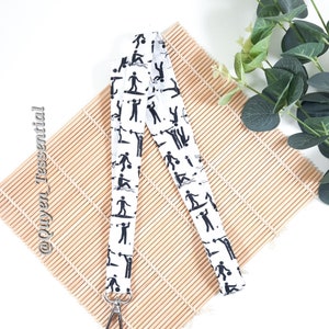 May include: White lanyard with a black and white pattern of stick figures playing different sports. The lanyard has a silver clip on one end.