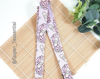 May include: A light purple lanyard with a repeating pattern of pink brains with bandages and small white flowers. The lanyard has a silver-colored metal clasp and is displayed on a bamboo mat.