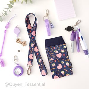 Könnte beinhalten: Eine Auswahl an Accessoires zum Thema Medizin, darunter ein Schlüsselband, eine Ausweisrolle, ein Stifthalter und ein kleiner Notizblock. Die Artikel haben einen marineblauen Hintergrund mit rosa Herzen, Blumen und Stethoskopen. Weitere Artikel sind ein lila Stift, eine kleine Uhr und ein Schlüsselanhänger.