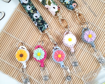 May include: A collection of floral patterned lanyards with matching daisy-shaped badge reels. The lanyards feature various floral designs on a dark background. The badge reels are in shades of yellow, pink, blue, and purple, each with a colorful daisy design.
