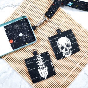 Könnte beinhalten: Drei schwarze Stoffschlüsselanhänger mit weißen anatomischen Illustrationen. Ein Schlüsselanhänger zeigt einen Schädel mit der Aufschrift "Nasal bone". Ein weiterer Schlüsselanhänger zeigt eine Wirbelsäule mit der Aufschrift "Unbar Vertebrae". Der dritte Schlüsselanhänger zeigt ein Weltraum-Design mit Sternen und Planeten.