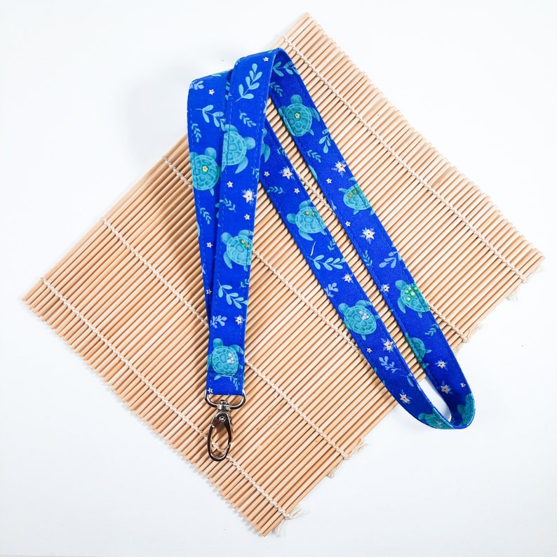 May include: A blue lanyard with a pattern of green sea turtles and white flowers. The lanyard has a silver metal clip.