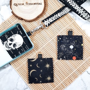 May include: Two black fabric keychains with different designs. One keychain features a celestial pattern with moons and stars, while the other features a galaxy pattern with planets and stars. Both keychains have a black ribbon loop and a gold metal ring.