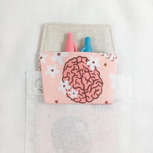 May include: A pink fabric pocket with a brain and flower print. The pocket is attached to a white shirt and has two pens inside.
