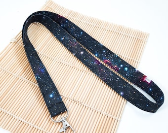 May include: A black lanyard with a galaxy print and a silver swivel clip. The lanyard is on a bamboo mat.