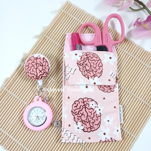 May include: A pink medical-themed set featuring a brain-patterned pocket organizer, a matching retractable badge reel with a watch, and pink scissors. The organizer holds a pen and other small tools. The set is decorated with brain and flower patterns.