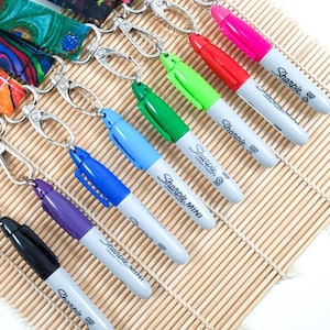 May include: A selection of Sharpie Mini markers in a range of colours, such as pink, red, green, blue, purple, and black, each connected to a silver keyring. The markers are displayed diagonally on a light brown bamboo mat.
