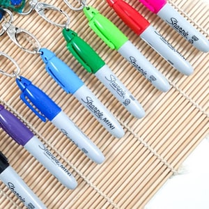 以下が含まれることがあります： さまざまな色のSharpie Miniマーカーのコレクション。各マーカーはシルバーのキーホルダーに取り付けられています。マーカーは、薄茶色の竹マットに斜めに配置されています。色は黒、紫、青、緑、赤、ピンクです。