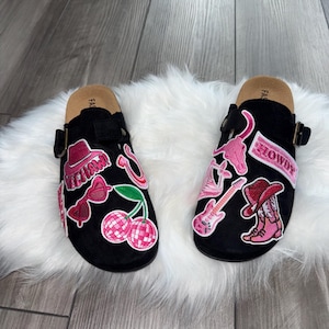 May include: Black clogs with a cork footbed and adjustable buckle. Each clog is decorated with pink and red patches, including a cowboy hat, cherries, and the word "HOWDY".