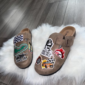 Könnte beinhalten: Ein Paar braune Wildleder-Clogs mit Aufnähern zum Thema Rennsport. Ein Clog zeigt eine Zielflagge, eine Rennstrecke und eine Autografik. Der andere Clog hat einen Aufnäher mit der Aufschrift "RAISE HELL PRAISE DALE" und eine Autografik.