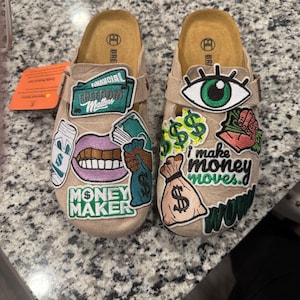 May include: A pair of beige clogs adorned with patches featuring money-related graphics and phrases. Patches include a green eye, dollar signs, a mouth, and text that says "Money Maker" and "I make money moves."