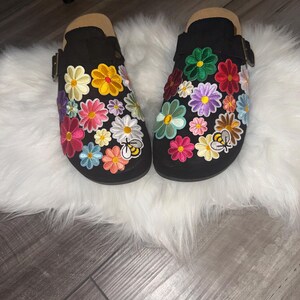 以下が含まれることがあります： ピンク、黄色、青、緑、白の色合いのカラフルな刺繍の花柄と、小さなハチの刺繍が施された黒いスリッポンタイプのクロッグ。各靴には、金属製のバックルが付いたサイドストラップが付いています。