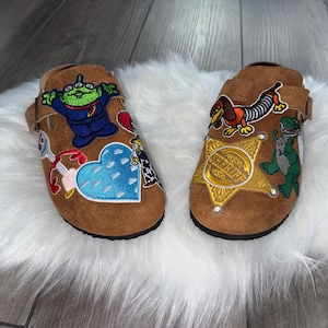 May include: A pair of brown clogs with various embroidered character patches. One clog features a green alien, a blue heart, and other designs. The other clog has a sheriff badge, a green dinosaur, and a dachshund.