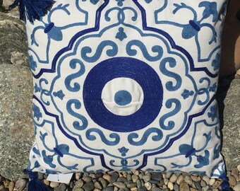 May include: Square white pillow with a blue embroidered medallion design. The pillow has a central circular pattern and corner tassels. The design features intricate scrollwork and floral motifs. The pillow is placed on a bed of small stones.
