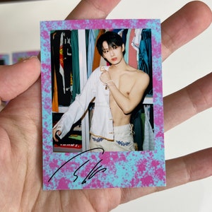 May include: A polaroid photo with a blue and pink splatter background. The photo shows a man with dark hair, wearing a white shirt, standing in a closet. The photo is signed with a black signature.