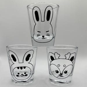May include: Three clear glass shot glasses with black and white cartoon animal designs. The glasses feature a bunny, a bunny wearing a hat, and a deer.