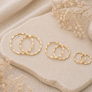 May include: Several pairs of gold-coloured, textured hoop earrings in different sizes. The earrings are displayed on a light-coloured, textured surface, with a neutral background and decorative sprigs.