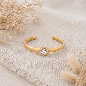May include: A gold open ring featuring a central, clear gemstone. The ring is marked with "IN 375" on the band. It's presented on a beige linen cloth, with dried flowers adding to the composition. The ring's design is minimalist.