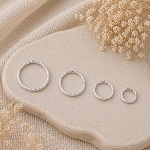 May include: Four silver-colored, textured hoop earrings of varying sizes are arranged on a light beige, oval-shaped stone. The earrings are displayed on a neutral-toned, textured fabric background, with dried flowers and a stone accent.