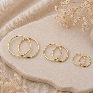 May include: Three pairs of gold hoop earrings in varying sizes are displayed on a light beige, textured surface. The earrings are arranged in a group, showcasing their circular design and shiny finish. The background features a soft, neutral fabric and delicate floral accents.