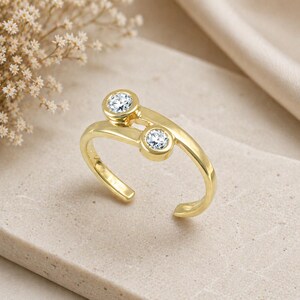 Pu&ograve; includere: Un anello color oro con due pietre preziose rotonde e trasparenti. L'anello ha un design aperto unico, con le pietre incastonate in castoni. L'anello &egrave; presentato su una superficie neutra, con uno sfondo morbido e sfocato.