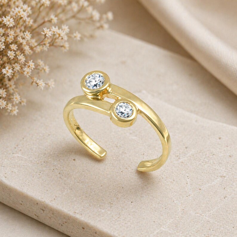 Pu&ograve; includere: Un anello color oro con due pietre preziose rotonde e trasparenti. L'anello ha un design aperto unico, con le pietre incastonate in castoni. L'anello &egrave; presentato su una superficie neutra, con uno sfondo morbido e sfocato.